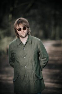 A person with shoulder-length hair and a beard stands outdoors, wearing sunglasses, a green button-up coat, and jeans. They have their hands behind their back and are looking forward with a neutral expression. The background is a blurred natural setting.