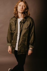 A person with long, wavy hair stands against a plain, dark background. They are wearing a green, military-style jacket over a white shirt and black pants, with bracelets on one wrist. The person's expression is neutral, and they are leaning slightly to one side.