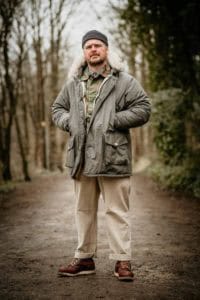 A man stands on a forest path, dressed in a gray parka with a fur-lined hood, a dark beanie, beige pants, and brown boots. He has a short beard and looks directly at the camera with a serious expression. The background features tall leafless trees and dense foliage.