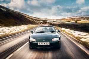 A small, dark green convertible sports car with the license plate "S77 KJU" is driving on a two-lane road through a hilly, partially snow-covered landscape. The driver is visible, and the weather appears clear with some clouds in the sky.
