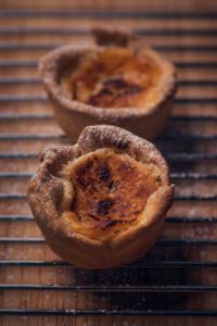 Two golden-brown, slightly burnt custard tarts rest on a wire cooling rack. The edges of the tarts are uneven and slightly flaky, showcasing the rustic appearance of the baked pastry. The surface of the custard has a caramelized texture.