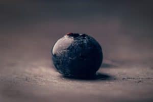 Close-up of a single blueberry on a textured surface, with a dark and moody background. The blueberry appears fresh with a slight sheen, highlighting its natural texture and color. The lighting emphasizes the berry's details, creating a dramatic effect.