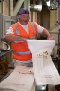 A man wearing an orange safety vest and a blue hair net smiles while holding a white plastic bag with the Muntons logo on it. He is standing in an industrial setting, likely a production or packaging facility.