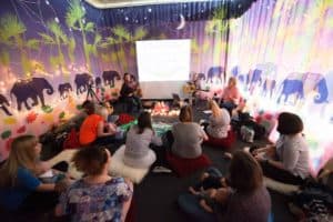 A group of people sit on colorful cushions in a vibrant room with a jungle-themed mural on the walls, featuring elephants. They are gathered around two musicians playing guitars in front of a projected screen, creating a warm and cozy atmosphere.