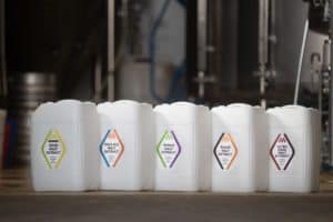 Five white plastic jugs, each labeled differently, stand side by side in a row. The labels indicate different types of malt extract: Sour, Pale Ale, Wheat, Roast, and Ultra Dark. The background is a blurred industrial setting with tanks and pipes.