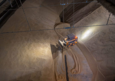 A top-down view of a large indoor grain storage facility with a skid-steer loader moving a mound of grain. The machinery has its headlights on, illuminating the grains and leaving tracks as it maneuvers through the space.