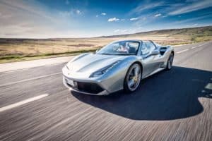 A silver ferrari convertible sports car driving on an open road through a scenic countryside on a clear day. The background features hills and a partly cloudy sky, creating a sense of speed and freedom—truly an image worthy of any automotive gallery.
