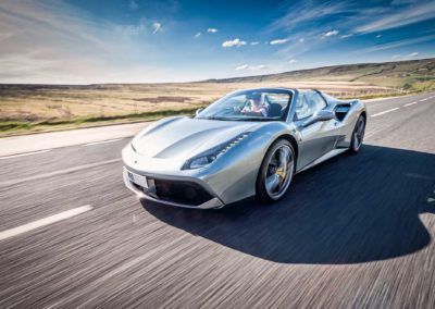 A silver ferrari convertible sports car driving on an open road through a scenic countryside on a clear day. The background features hills and a partly cloudy sky, creating a sense of speed and freedom—truly an image worthy of any automotive gallery.