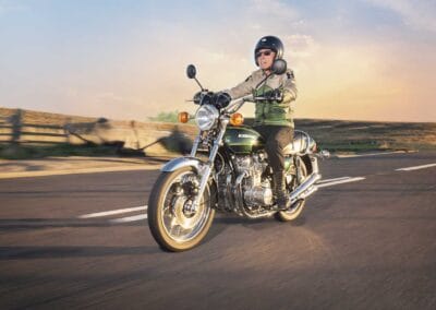 A person wearing a helmet and sunglasses rides a classic kawasaki z 900 motorcycle on an open road during sunset, capturing the spirit of an automotive gallery. The scenery includes a clear sky and distant fields, creating a sense of freedom and adventure.