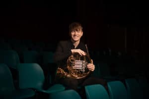 A young man in a dark outfit sits alone in a dimly lit theater. He smiles at the camera while holding a French horn. The teal-colored seats around him are empty, and the background is shadowy, capturing the essence of brand photographs in Manchester.