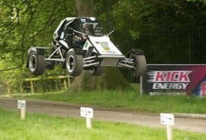 A dune buggy is airborne during a high-speed jump on a dirt path through a park with trees in the background. The buggy has a white and black design and displays a visible sponsor logo. A "KICK Energy" banner is placed in the background near the path.