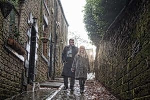 A man and a woman walk arm in arm down a narrow, cobblestone alley lined with stone walls and buildings. The man wears a coat and scarf, the woman a patterned coat. A cat follows behind them. They both appear to be engaged in conversation, smiling.
