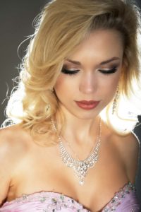 A woman with blonde, wavy hair and makeup is looking down. She is wearing a strapless pink dress adorned with rhinestones and a sparkling necklace and earrings. The light highlights her hair and face, creating a soft glow.