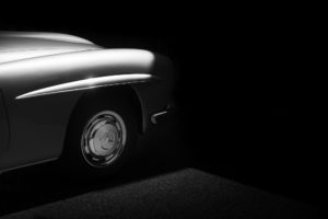 A classic car's front end is partially illuminated, revealing one headlight, a chrome-finished wheel, and a section of the fender against a dark background. The car's curves and details are highlighted by a soft light, creating a dramatic and vintage aesthetic.