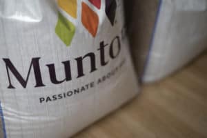 Close-up of a sack with the word "Munto" and the slogan "Passionate About You" written on it. A colorful diamond-shaped pattern is visible above the text. The sack, placed on a wooden surface, is part of brand photographs captured in Manchester, with another similar sack partially visible in the background.