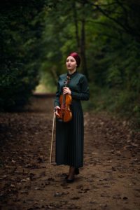 A woman with a red headscarf and a long dark green dress stands on a forest path, holding a violin and bow. The path is covered with fallen leaves and surrounded by lush, green trees. The lighting is soft, creating a serene and peaceful atmosphere.