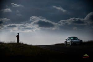 A silhouette of a person standing on a grassy hill, looking towards a silver sports car parked on a road. The sky is overcast with dark, dramatic clouds. The scene has a contemplative, cinematic feel. Photography credit is visible.