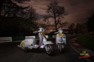 Two vintage scooters with racing numbers 99 and 991 are parked on a dimly lit road surrounded by leafless trees under a cloudy evening sky. The scooters are adorned with decals, and the scene is set in a wooded area. The photographer's credit is visible in the bottom right.