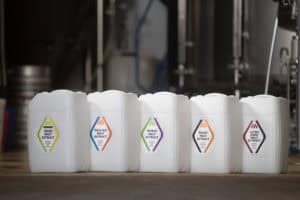 Five white containers with labels in various colors, each displaying different types of malt extract: Sour Malt Extract, Pale Ale Malt Extract, Wheat Malt Extract, Roast Malt Extract, and Ultra Dark Malt Extract. Captured in a brewery setting with a hint of Brand Photographs Manchester's signature style.