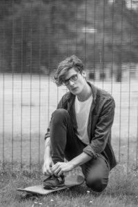 Black and white photo of a young person with glasses, crouching on the grass and tying their shoe. They are wearing a casual outfit with a t-shirt and open shirt, with a skateboard on the ground beside them. A wire fence is visible in the background.