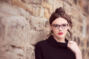 A woman with dark hair in a messy bun and wearing glasses and red lipstick stands against a textured stone wall. She is dressed in a black turtleneck sweater and looks directly at the camera with a neutral expression.