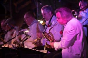 A jazz band performs on stage. Musicians, all dressed in white jackets, play various saxophones. The lighting on stage is dim with a purple hue, creating a vibrant atmosphere.