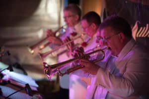 Three musicians in white suits play trumpets in unison during a live performance. They are closely lined up, focusing on their instruments, with music stands visible in front of them and stage lighting creating a warm atmosphere.
