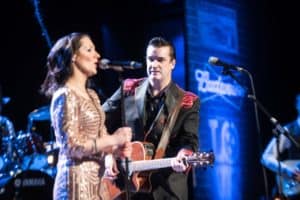 A woman in a sequined dress and a man in a suit with a guitar perform on stage with microphones in hand. The man is looking at the woman while playing guitar. The background displays blurred signs and musical instruments.