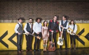 A seven-member band posing for a photo against a yellow and black wall. They are dressed in a mix of formal and casual attire, holding various musical instruments including guitars and a bass. The members are smiling and standing in a relaxed manner.