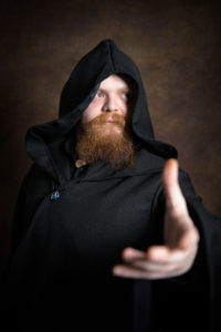 A bearded person dressed in a dark, hooded cloak extends their hand forward. The background is a dim, textured backdrop, adding to the mysterious and dramatic atmosphere of the image.