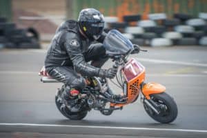 A rider dressed in black protective gear and a helmet races an orange mini motorcycle on a track. Tires line the background, and the rider's number, 71, is displayed on the front of the bike.