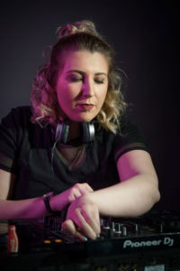 A woman with curly hair tied back, wearing headphones around her neck, is focusing intently while adjusting audio equipment on a DJ mixer. She is dressed in a black top with sheer sleeves, and the background is dark with subtle pink and purple lighting.