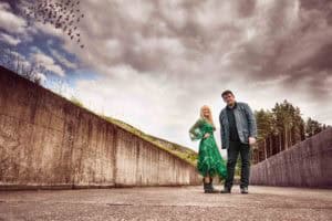 A woman in a green dress and a man in dark clothing stand together on a wide concrete path under a cloudy sky. Birds are flying in a V formation in the distance, and trees line the edge of the path. The atmosphere appears overcast and dramatic.
