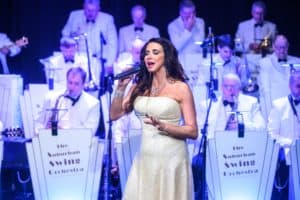 A woman in a sparkling white gown sings into a microphone on stage. Behind her, a big band orchestra, dressed in matching white suits and black bow ties, is playing instruments. The band’s music stands read "The Suburban Swing Orchestra." The stage is lit with blue lights.