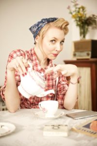 A woman with blonde hair styled in vintage curls, wearing a blue bandana and red plaid shirt, is seated at a table pouring tea from a floral teapot into a teacup. The setting has a retro feel with an old radio and flowers in the background.