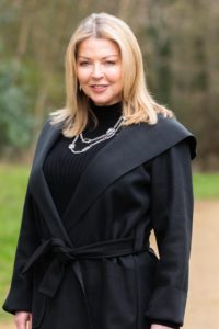 A woman with blonde hair, wearing a black coat and a layered necklace, stands outdoors on a pathway with blurred green foliage in the background. She smiles at the camera.