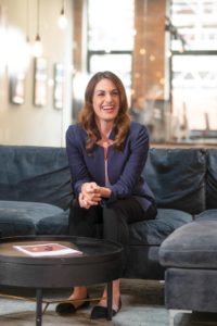 A smiling woman with long brown hair wearing a blue blazer and black pants sits on a dark grey couch in a modern office space. A black coffee table with a magazine on it is in front of her, and there are hanging lights and large windows in the background.