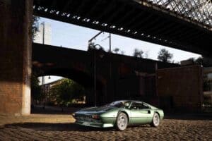 A classic green ferrari 308 sports car is elegantly parked on a cobblestone road under a large bridge, resembling an exhibit in an automotive gallery. The urban scene, with its mix of modern and brick buildings, basks in sunlight that casts playful shadows on the path.