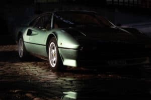 A classic green sports car, showcased like a masterpiece in an automotive gallery, is parked on a cobblestone street, partially illuminated by a light source. The image emphasizes its sleek design and vintage appeal, with reflections glistening on the wet cobblestones.
