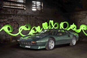 A vintage green sports car is parked inside an automotive gallery with industrial-style brick walls. Bright green light trails artistically swirl around the car, creating an eye-catching effect in the dimly lit setting.