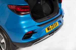 Close-up of the rear of a blue MG ZS electric vehicle with the trunk open. Photographing the brand new MG, the image showcases the vehicle's taillight design, rear bumper, and a license plate with the text "MG ZS EV." The car is set against a white background.