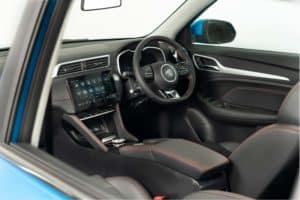 The image shows the interior of a modern car featuring a black, red-stitched steering wheel, a digital dashboard, a touchscreen infotainment system, and leather seats. Photographing the brand new MG reveals its sleek and technologically advanced appearance with a blue exterior visible through the window.