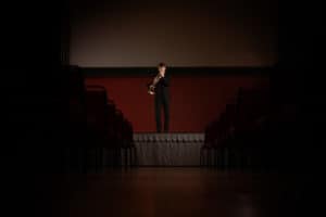 A musician stands on a stage, playing a brass instrument, in a dimly lit auditorium. The stage is illuminated while the surrounding audience area remains dark. Rows of empty chairs are visible on either side of the frame, leading up to the performer.