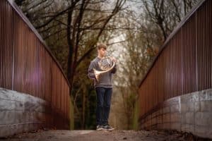 A young person stands on a wooden pathway in a forest holding a French horn. They are dressed casually in jeans and a sweater. The trees in the background are barren, suggesting autumn or winter. The pathway is lined with wooden beams on either side.