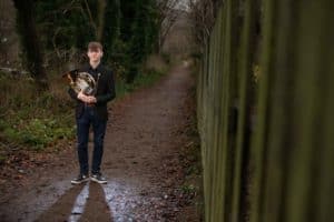 A young man stands on a wooded path holding a French horn. He is wearing a black jacket, dark pants, and sneakers. The path is bordered by trees with dense foliage, and a wooden fence is visible on the right side of the image. The ground is wet and covered with leaves.