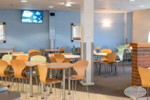 A spacious, well-lit cafeteria with a mix of wooden and metal chairs around tables. The walls are painted light blue, and a television screen displays content on the far wall. There are no people present in the room. The logo at the bottom right reads "Tim Simpson Architectural Photography Manchester.
