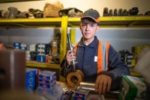 A young person wearing an orange safety vest and cap holds a machine part in an organized workshop. Shelves filled with boxes and tools are visible in the background. Captured by the UK's best commercial photographer, the individual looks focused amid various mechanical components.