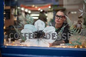 A person wearing black-framed glasses paints a large white flower with green leaves on a glass window from the inside of a cozy café. The words "COFFEE" and "DESSERT" are visible through the glass. The warmly lit scene feels like an artist's photoshoot, capturing creativity in action.