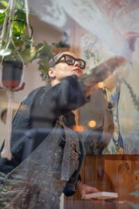 A person in glasses and overalls is painting a colorful mural on a wall, surrounded by hanging plants and various decorations. The scene, which appears to be an artist photoshoot, is viewed through a glass window.