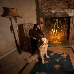 A person sits in an armchair next to a golden retriever, facing a fireplace with illuminated, stylized flames reminiscent of light painting photography. The setting is a rustic, dimly lit room with a worn-out carpet and an old-fashioned lamp.